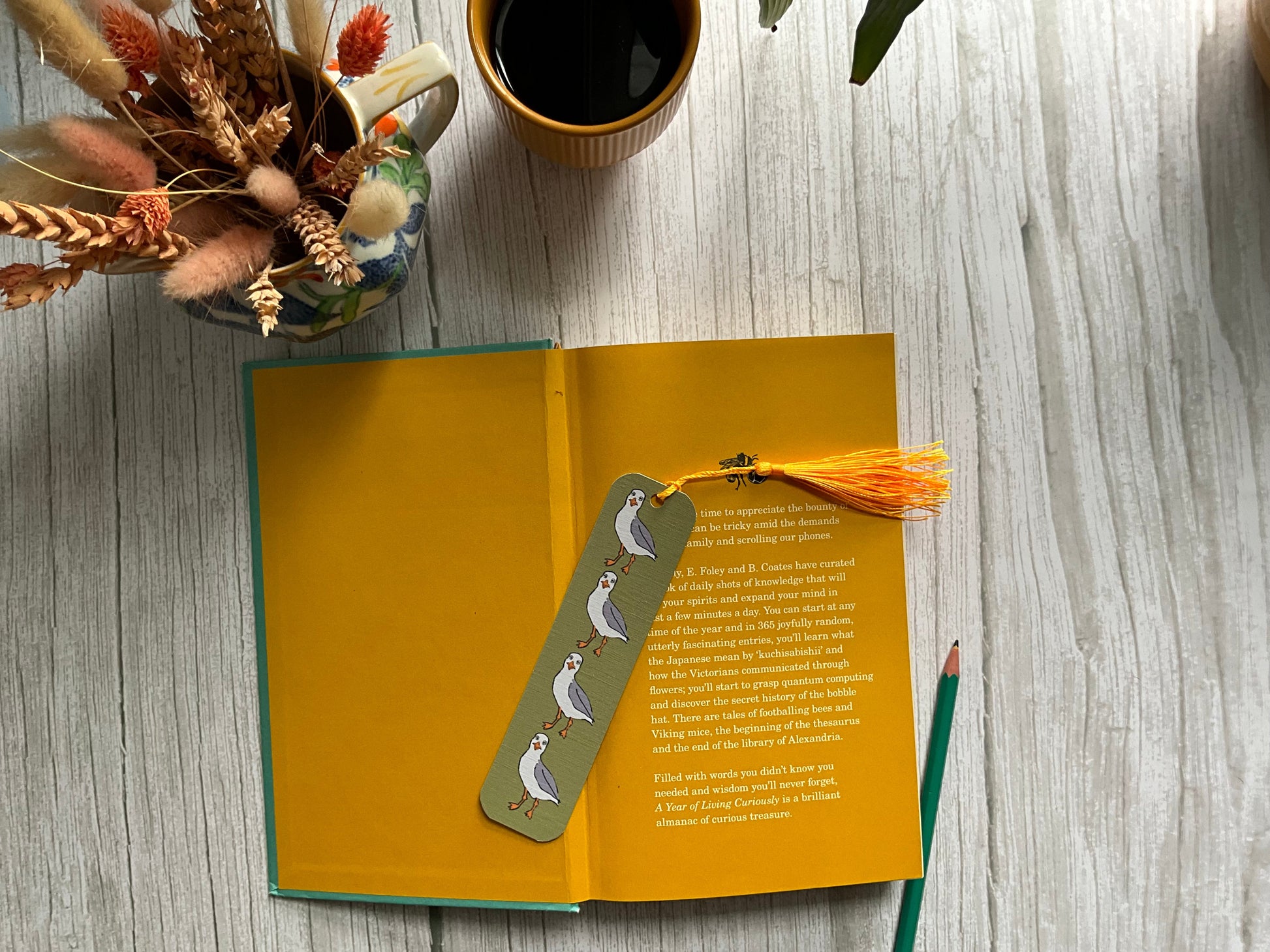 Open book with a green bookmark featuring hand drawn seagulls and an orange tassel on a wooden surface with a cup of coffee and dried flowers.
