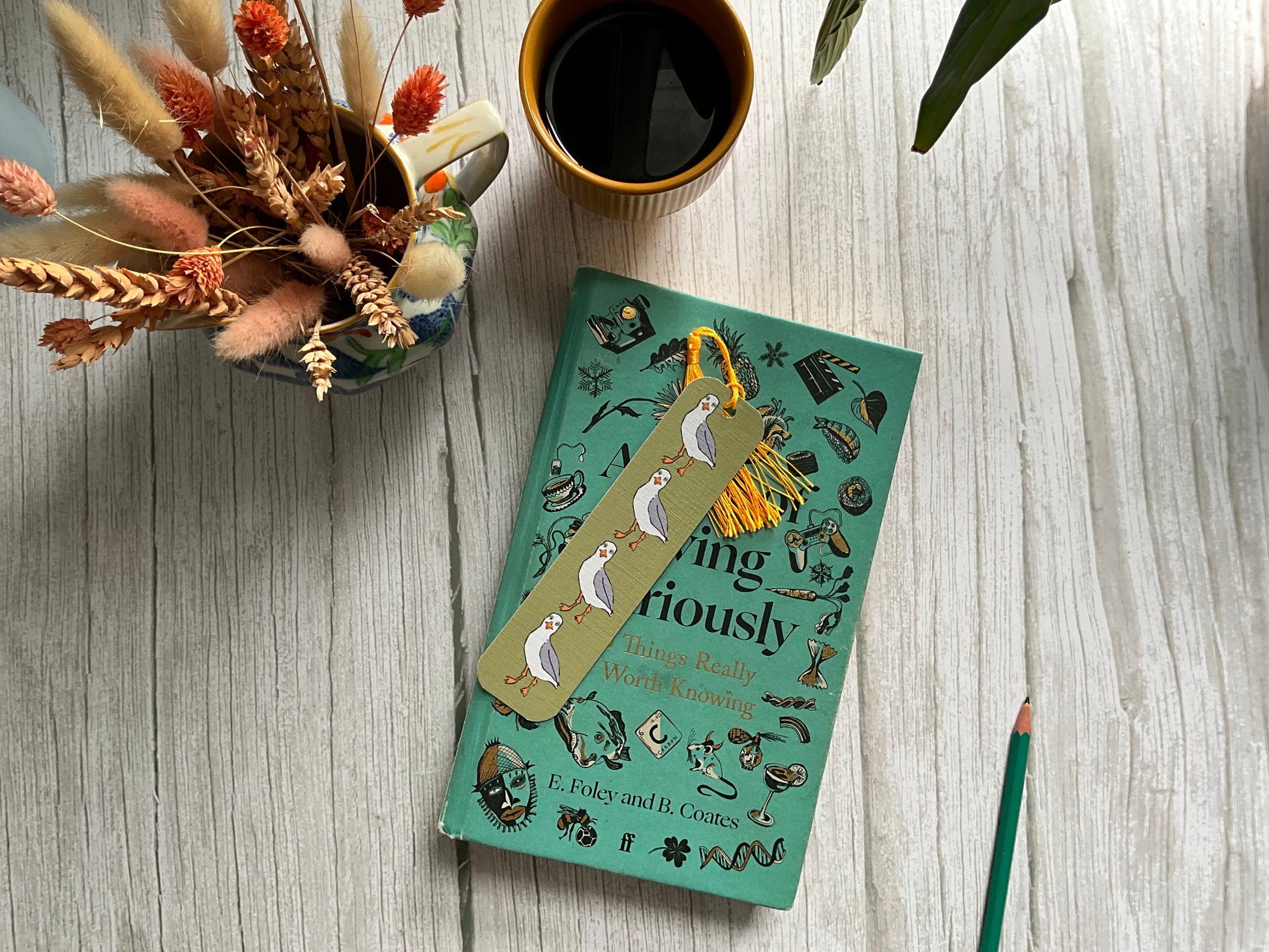 Green bookmark with seagull print pattern on a patterned book placed on a wooden surface with a cup of coffee and jug of fried flowers.