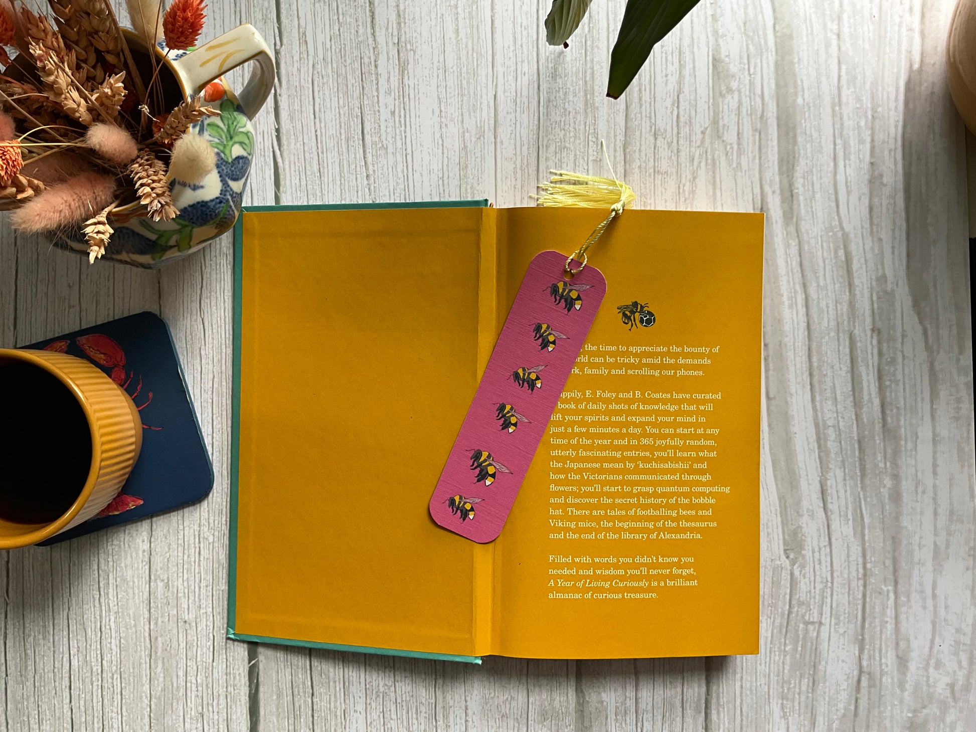 Open book with a decorative pink bookmark on a wooden surface, next to a cup and plant.