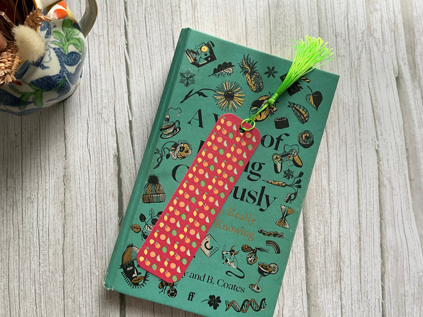 Book with a red citrus fruit printed bookmark with a bright green tassel. The book is on a wooden table with a vase of dried flowers 