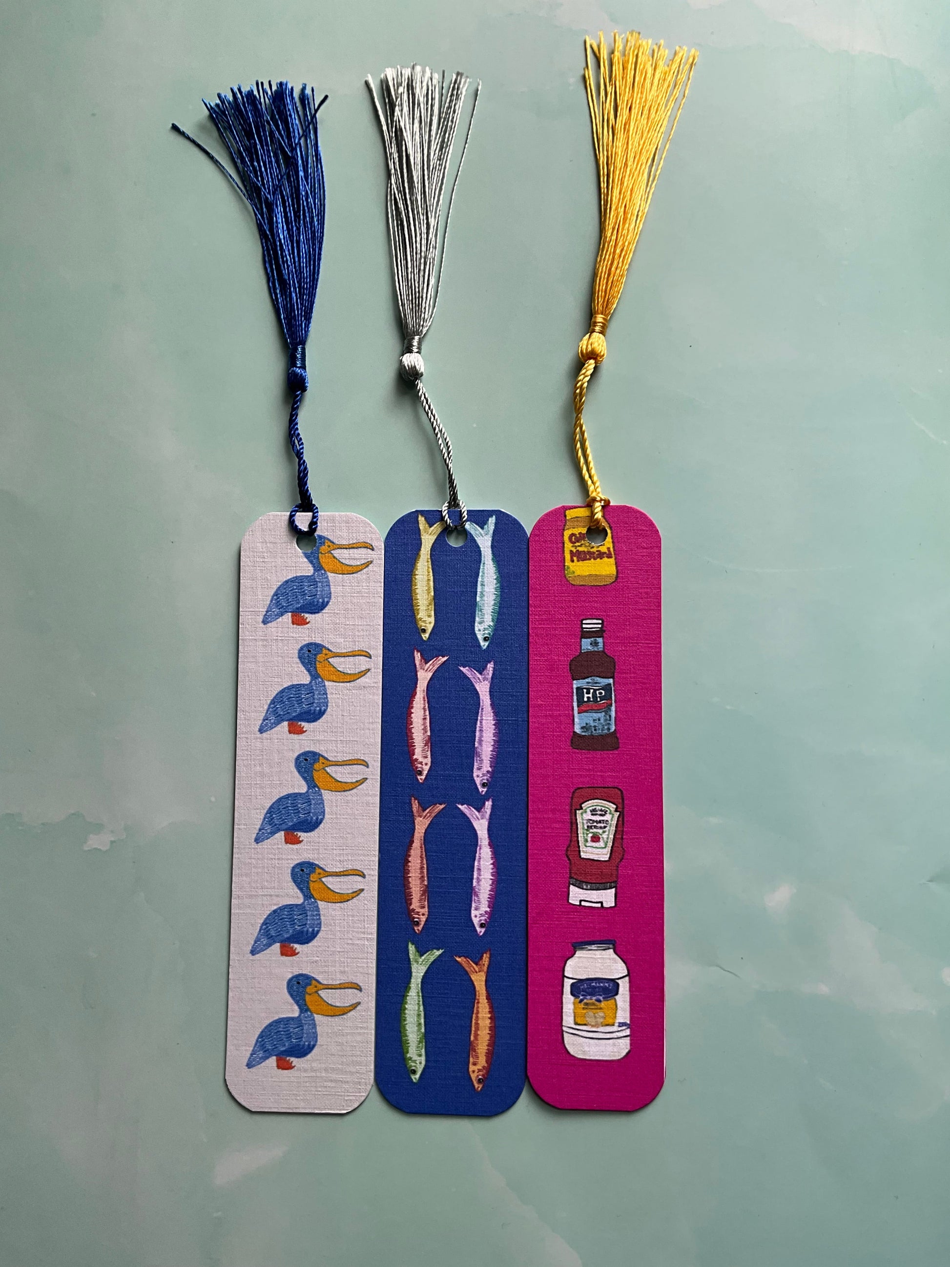 Three colorful bookmarks one featuring a bird print, one a fish print, and one a condiments print, all with tassels resting on a light blue background