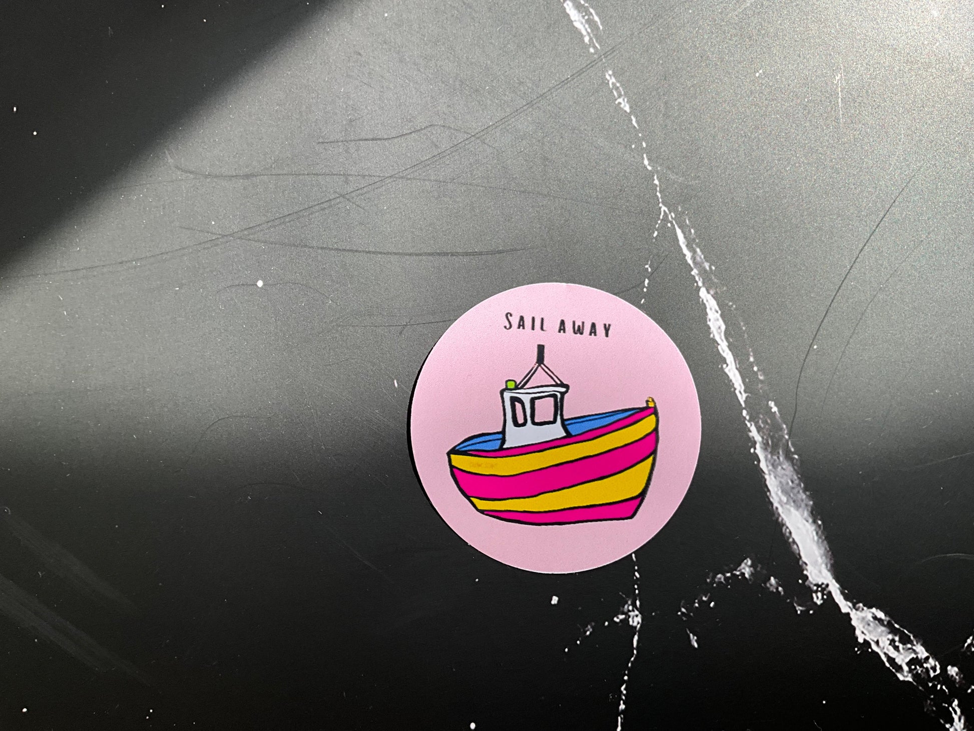 Sticker with a colorful boat design and 'Sail Away' text on a textured black surface.