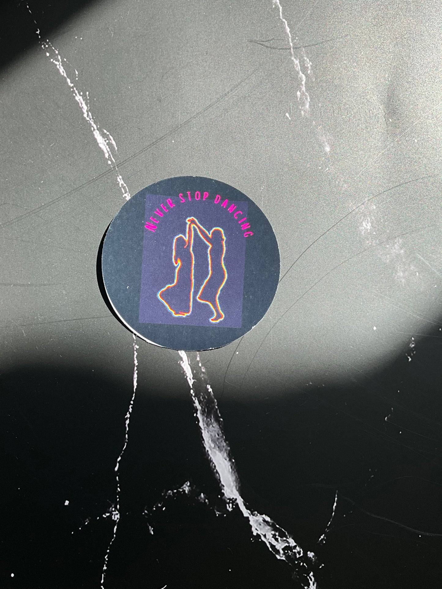 Circular black vinyl sticker with a rainbow coloured outline of two women dancing and the text ‘never stop dancing’ in pink letters. This sticker is on a sunny black marble surface. 