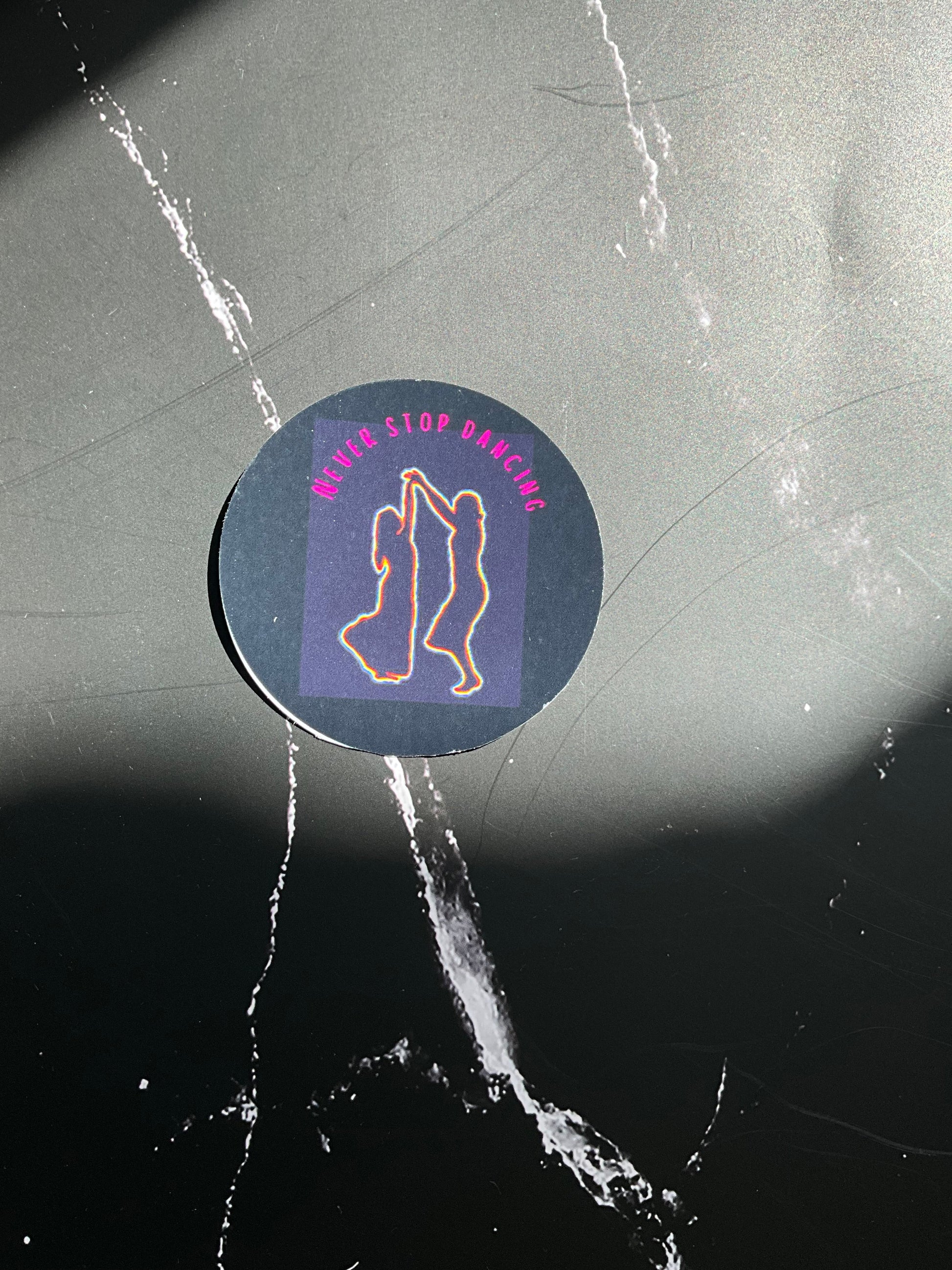 Circular black vinyl sticker with a rainbow coloured outline of two women dancing and the text ‘never stop dancing’ in pink letters. This sticker is on a sunny black marble surface. 