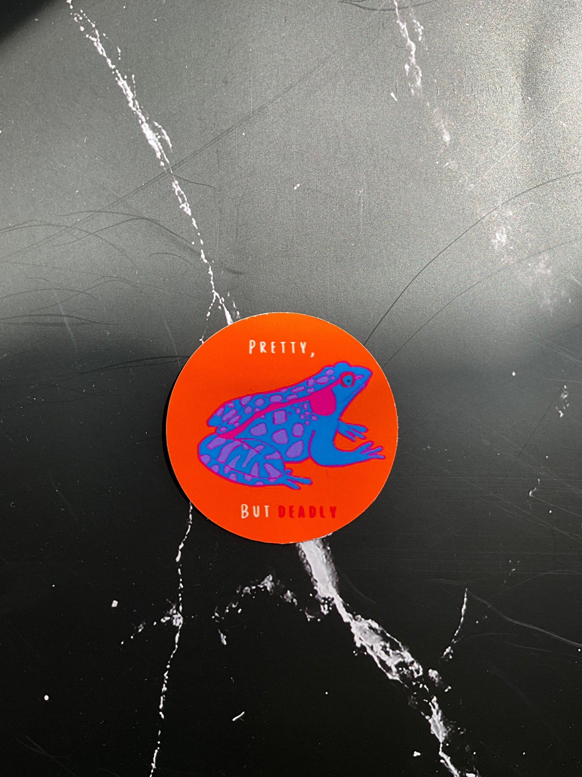 Round orange sticker with blue and pink graphic frog on a black marbled background