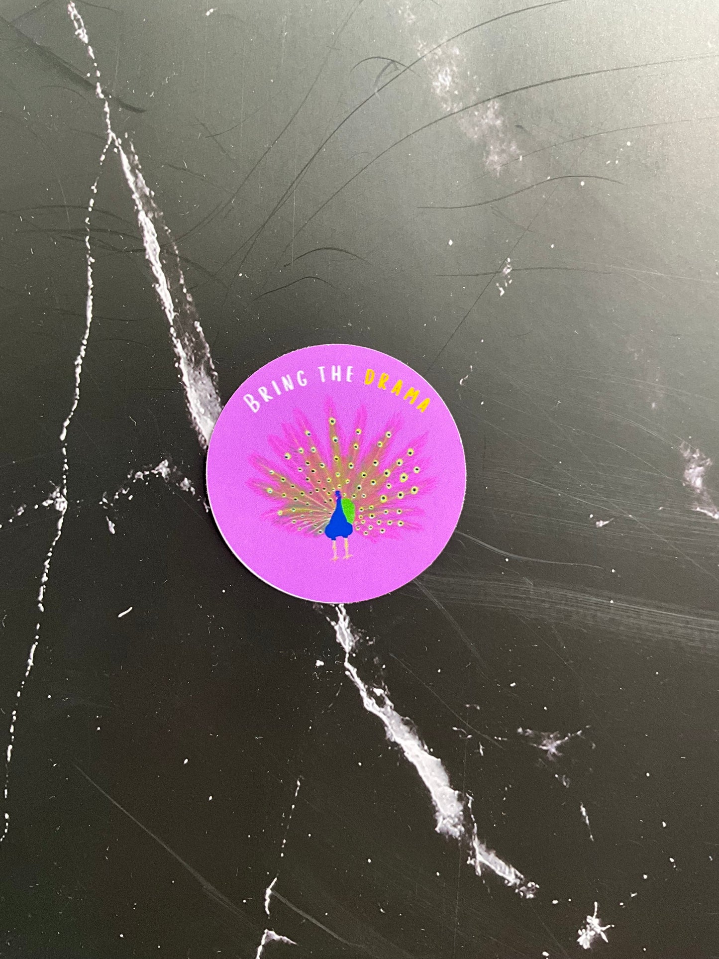Purple circular sticker with peacock design on a black marbled surface