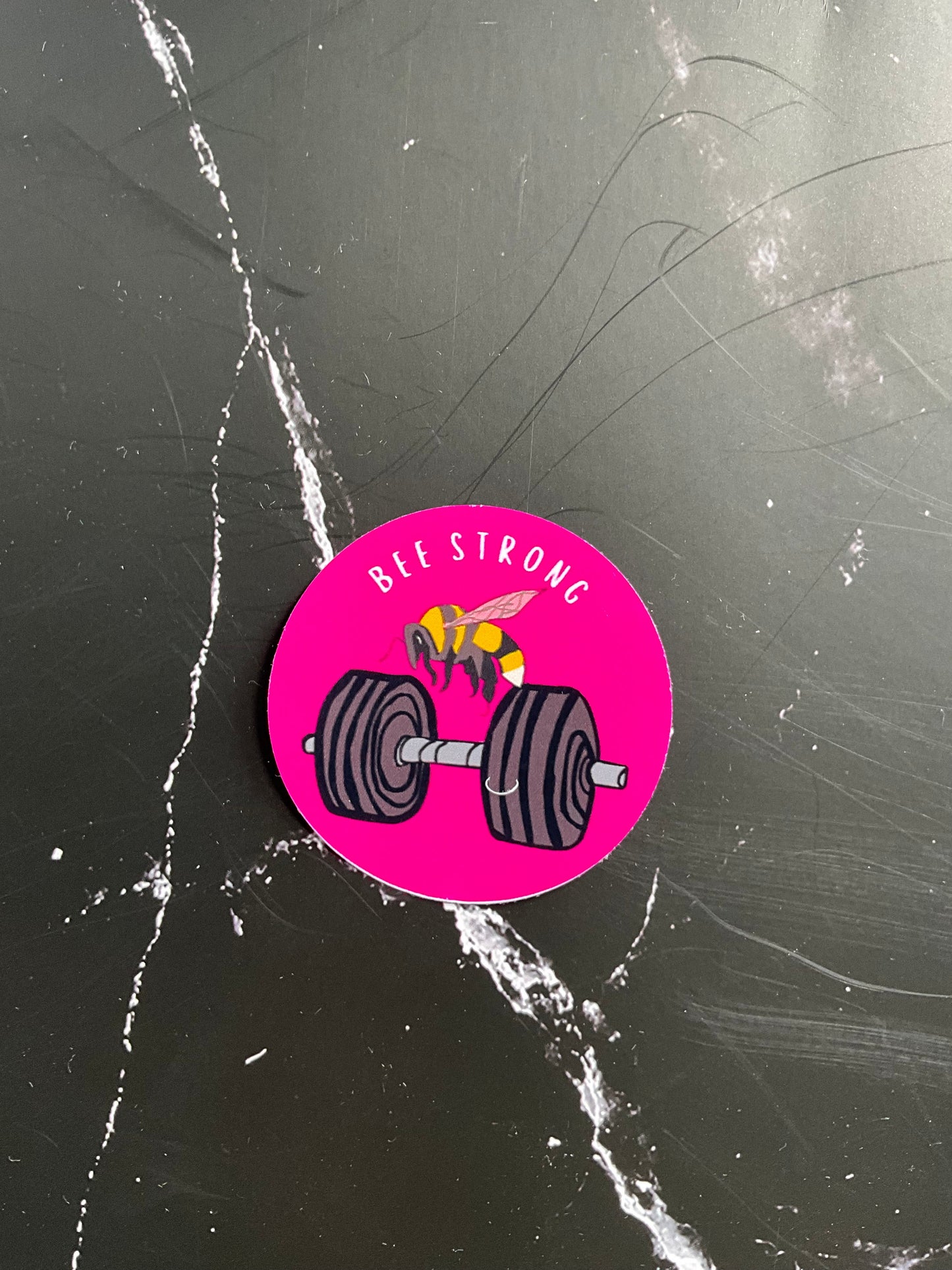 Pink circular sticker with a dumbbell  and bee graphic and text “bee strong” on a black surface