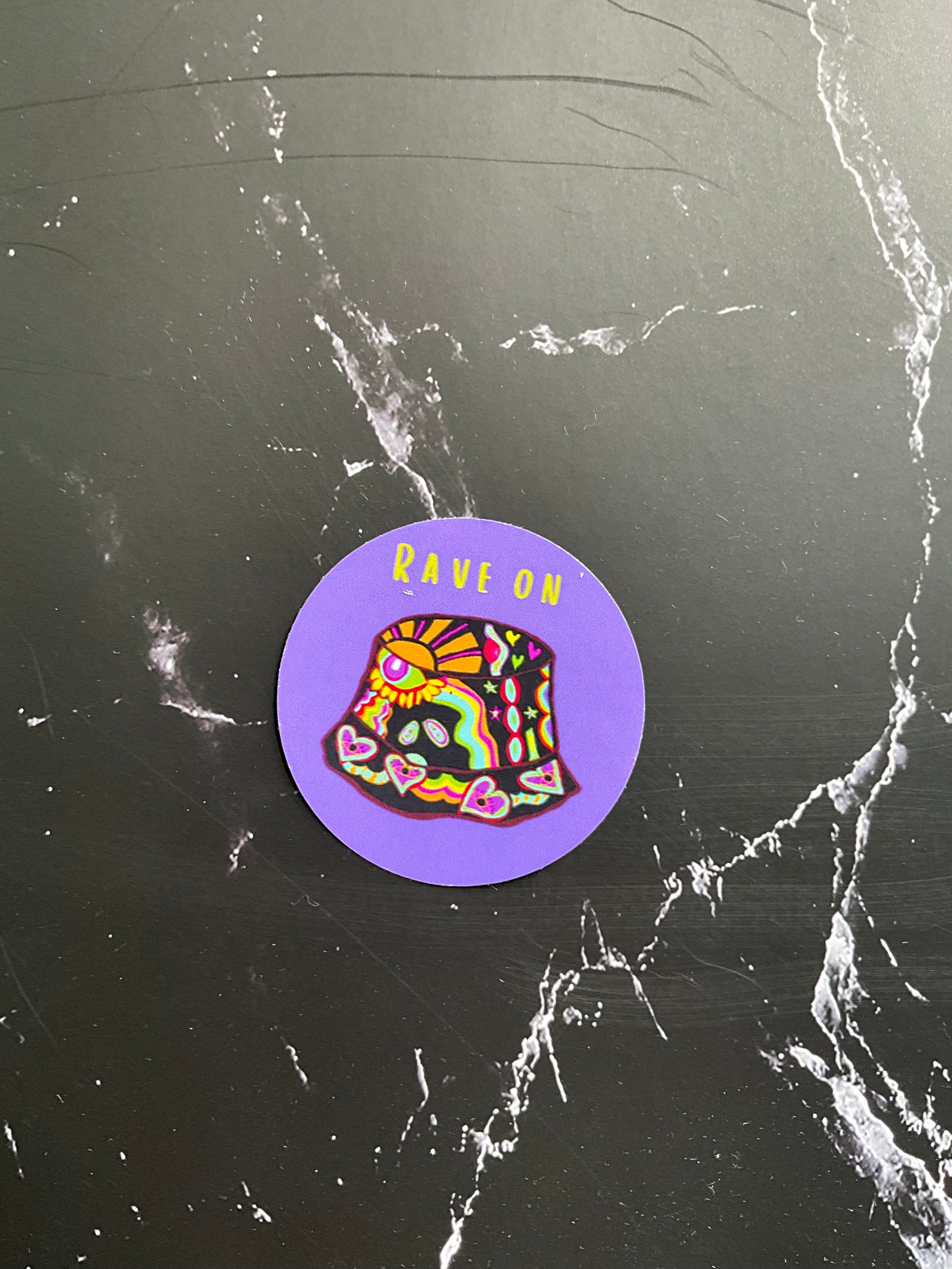 Retro round vinyl sticker featuring a colourful bucket hat and the words “rave on” on a black marble background 