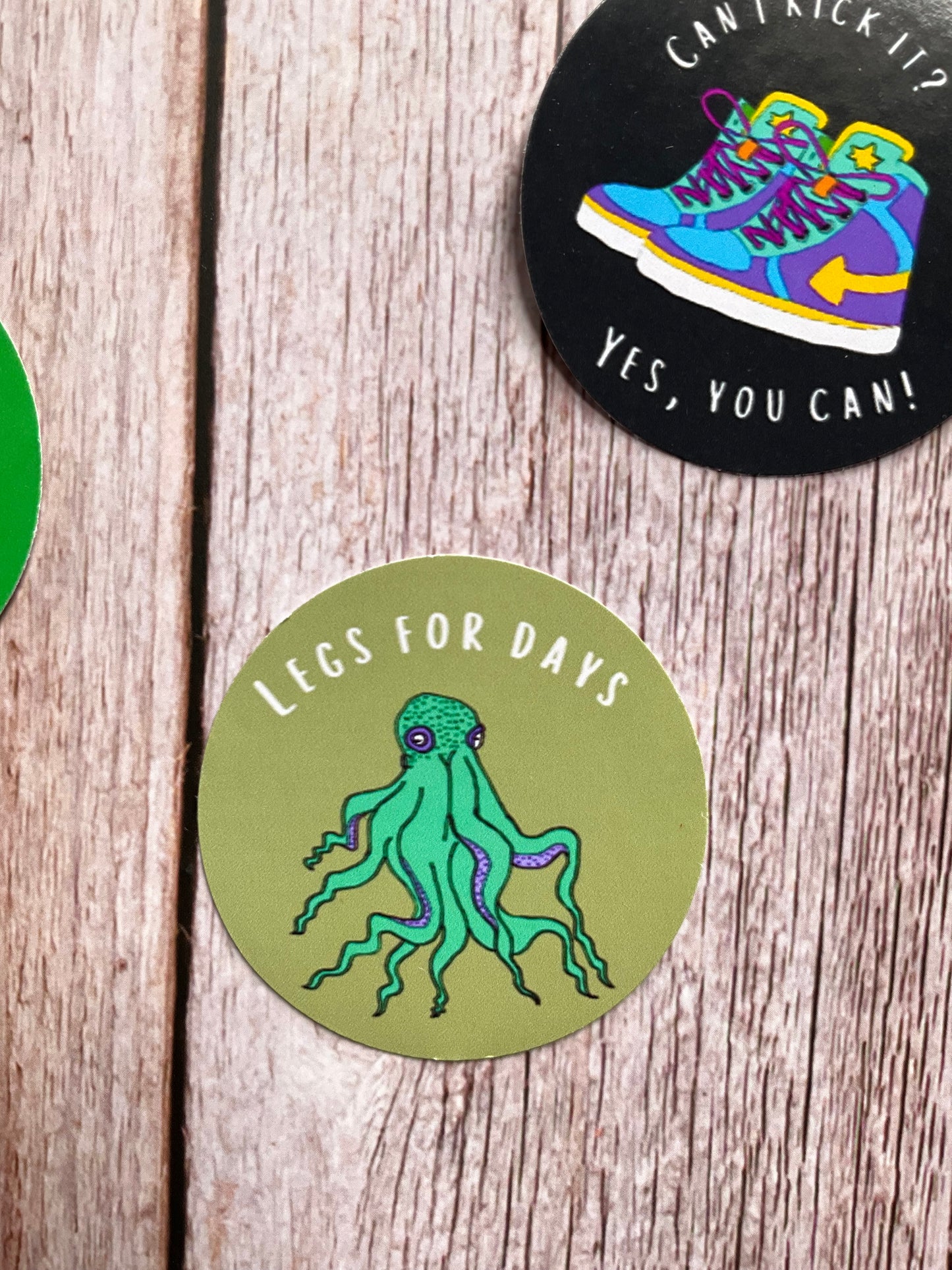 Two round stickers on a wooden surface, one with a colorful shoe design and text, the other with a green octopus illustration.