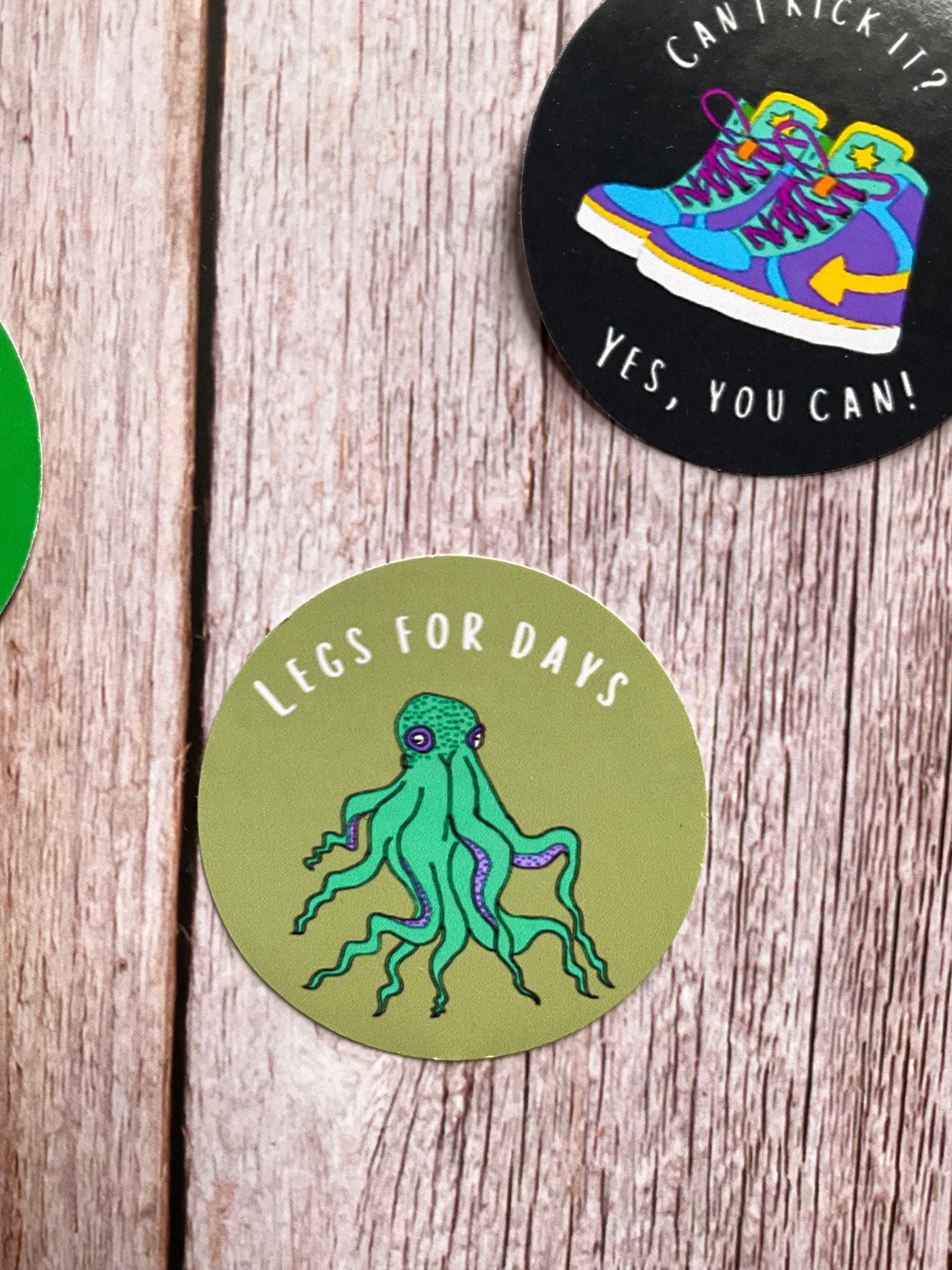 Two round stickers on a wooden surface, one with a colorful shoe design and text, the other with a green octopus illustration.