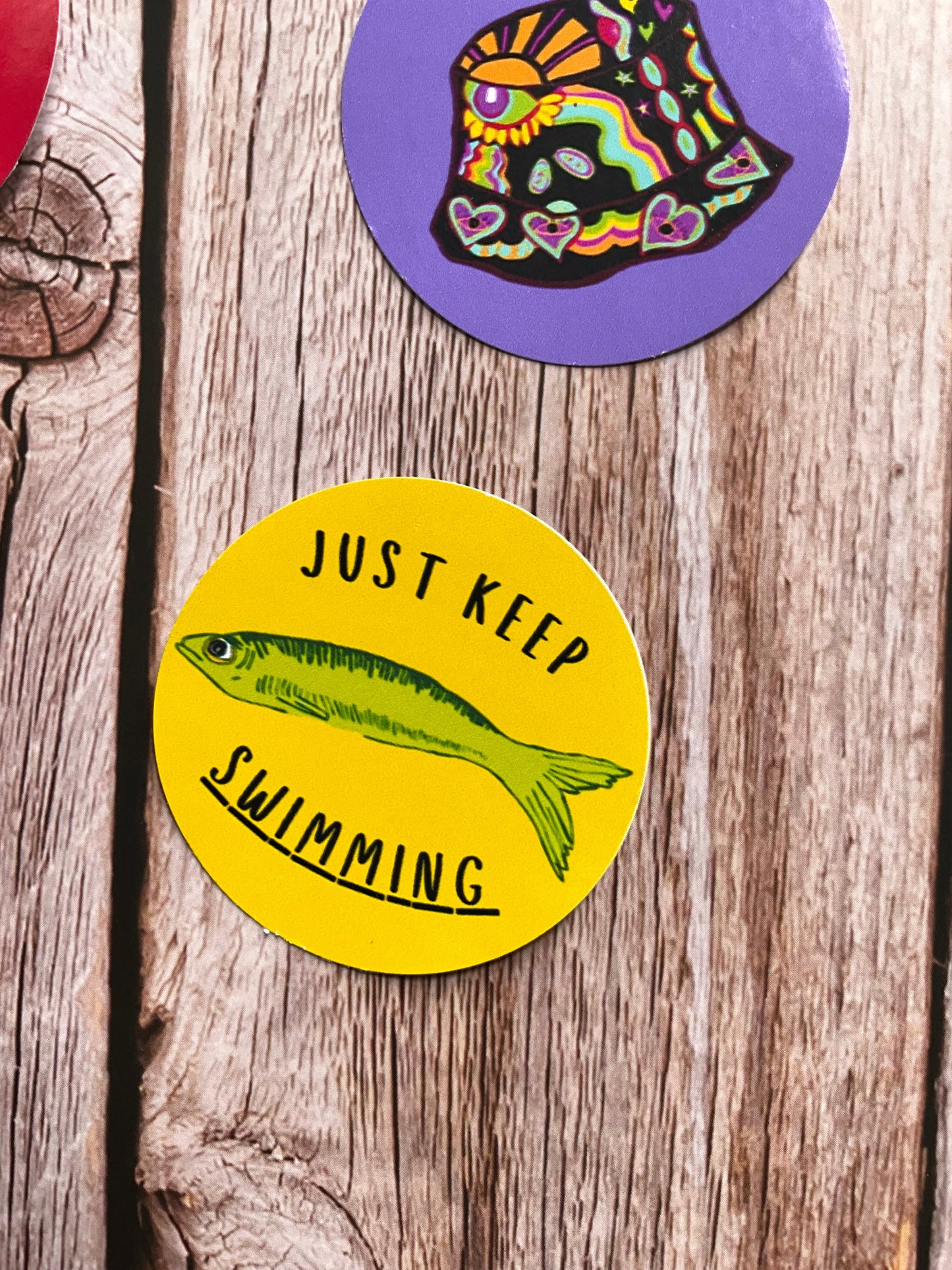 Round stickers on a wooden surface, one with a colourful fish design and with 'Just Keep Swimming' text.