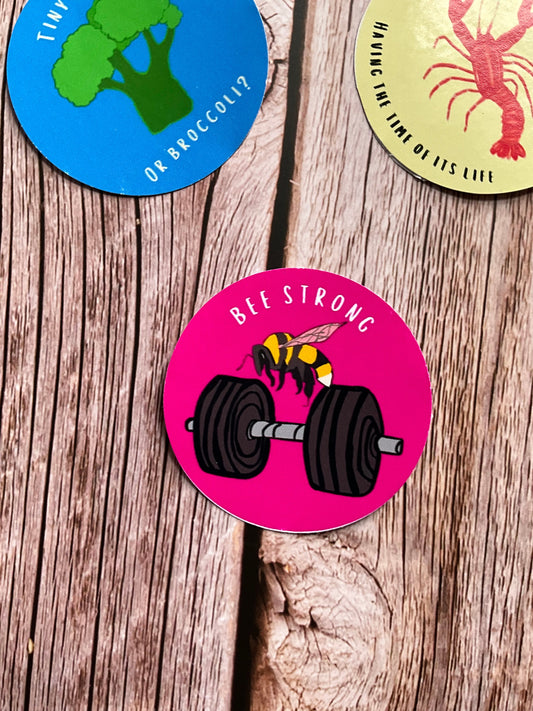 Vinyl stickers on a wooden surface - one featuring a bee & dumbbell illustration with the phrase ‘bee strong’