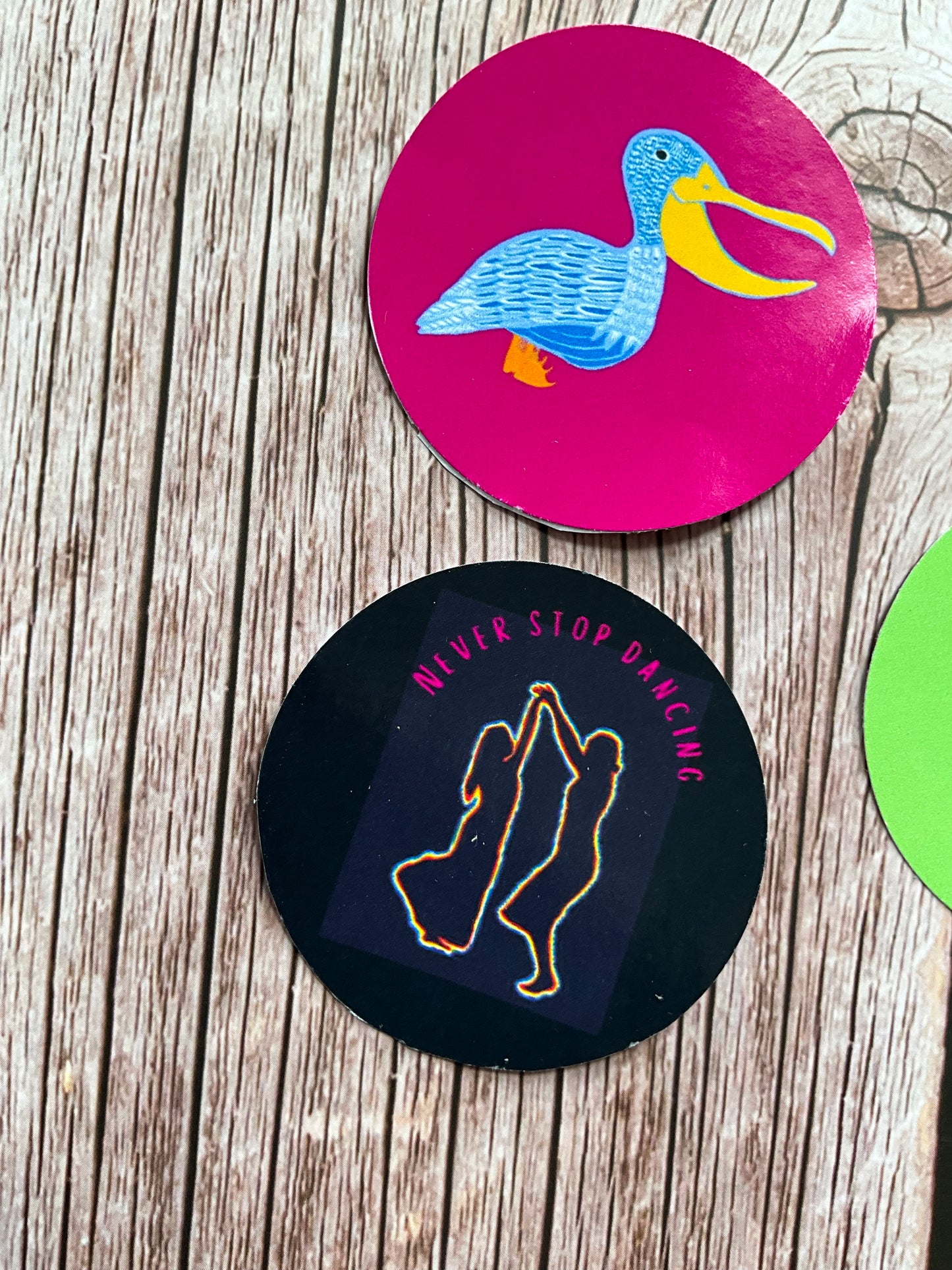 Two round stickers on a wooden surface, one with a blue and pink pelican design and the other with outlines of people dancing and the text ‘ never stop dancing’.