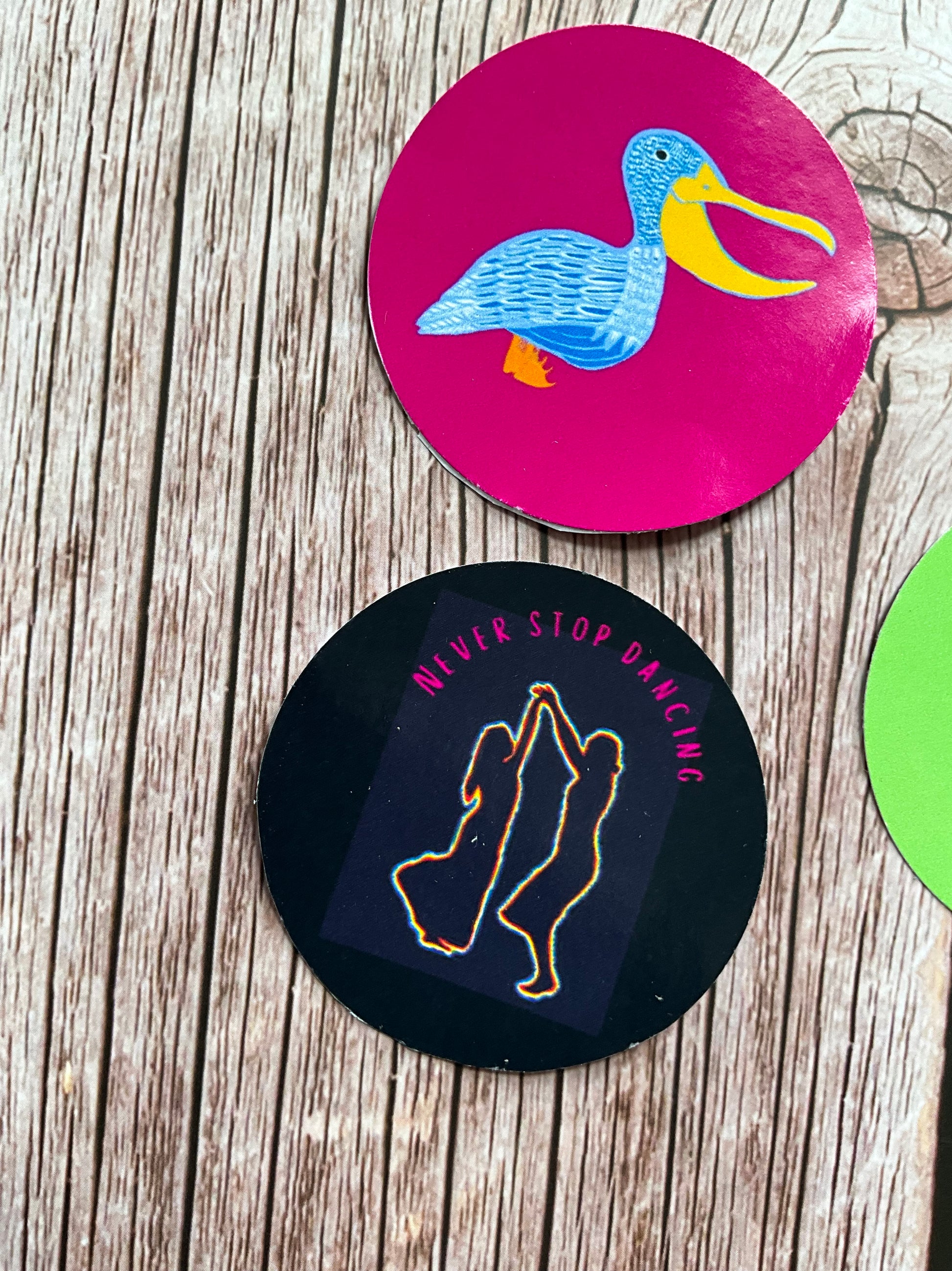 Two round stickers on a wooden surface, one with a blue and pink pelican design and the other with outlines of people dancing and the text ‘ never stop dancing’.
