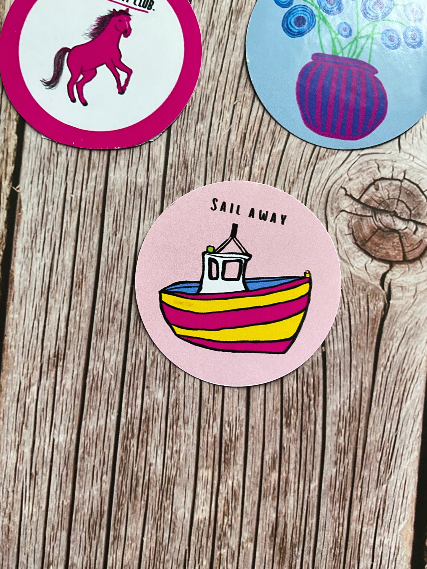 Round vinyl sticker featuring a colourful pink and yellow boat and the words ‘sail away’ in black letters.