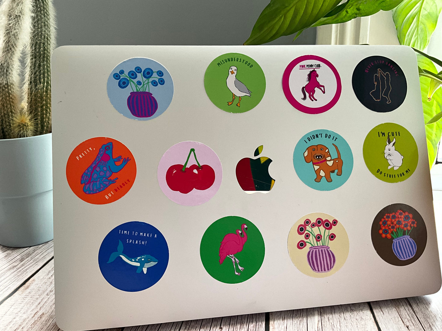Laptop with various colorful stickers on a wooden surface with plants in the background