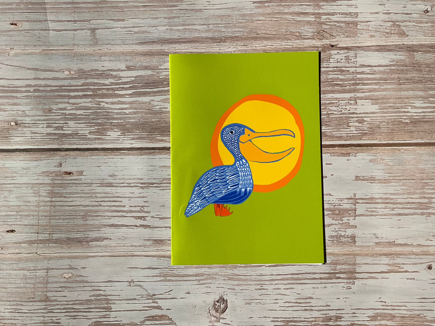 Card with a blue patterned pelican against a yelllow and orange sun. he card background is a lime-green colour. 