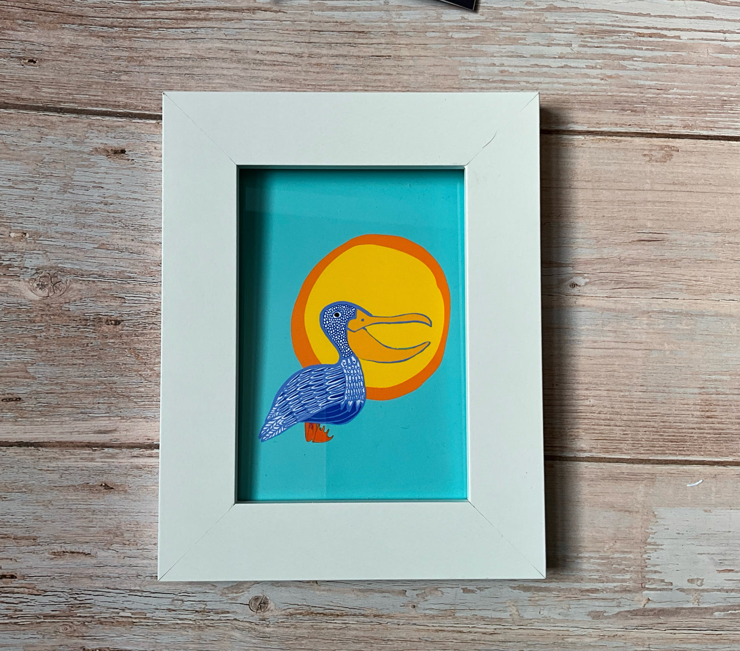 Playful bird print - patterned pelican in front of a large orange sun against a sky-blue background. Framed in a white wooden frame, set against a wooden background. 