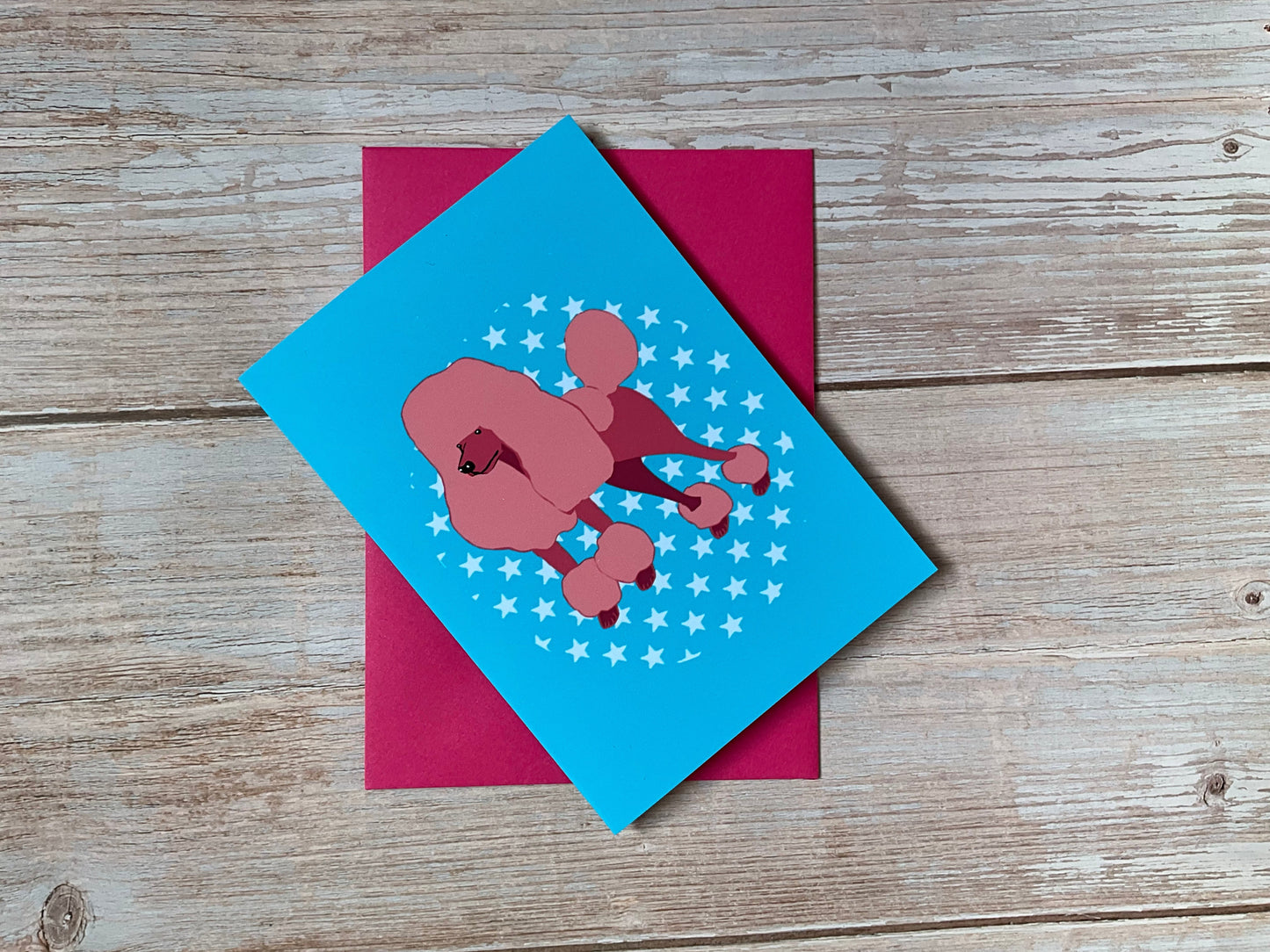 Blue card with pink poodle and stars design on a pink envelope against a wooden background