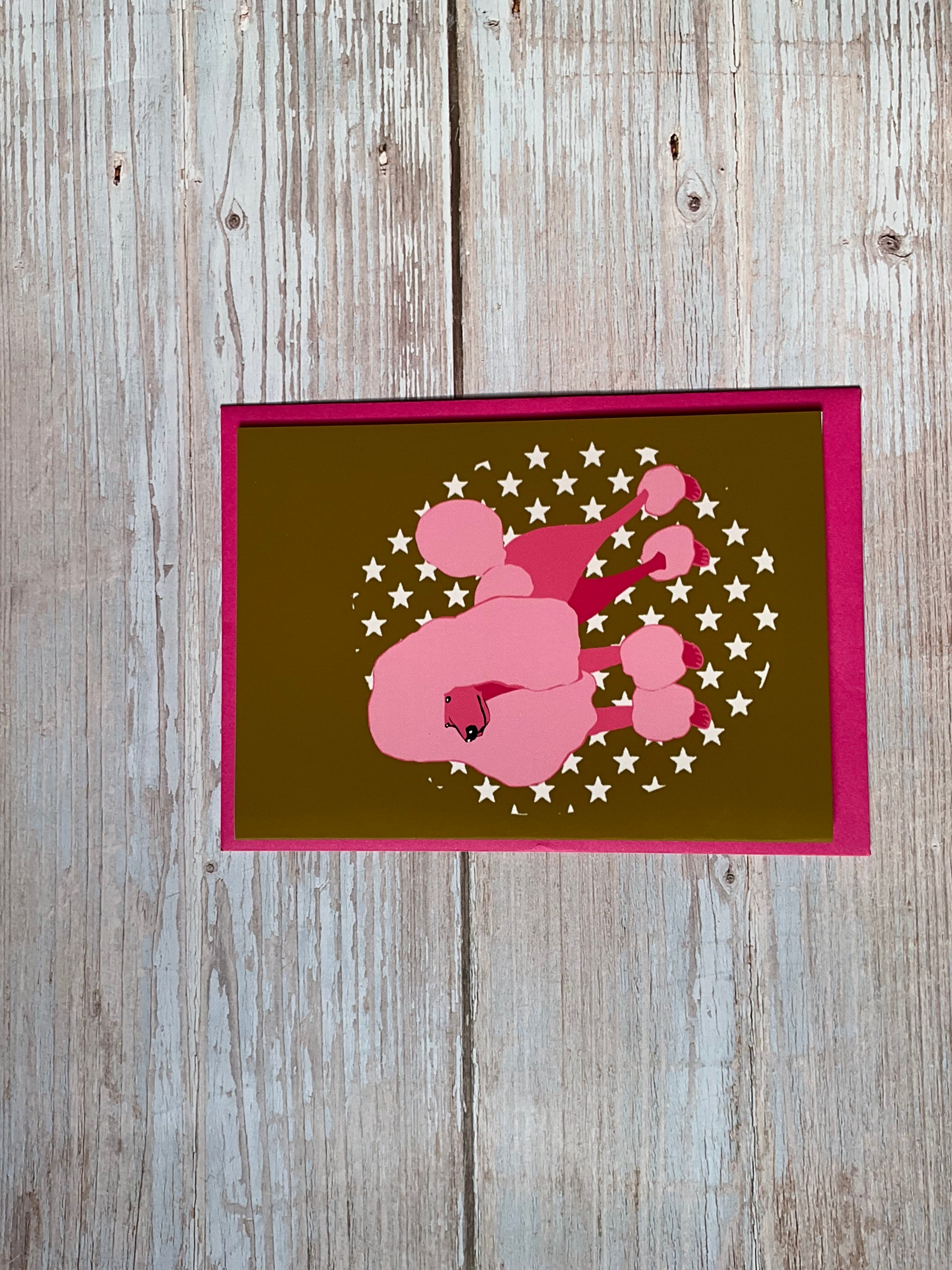 Greetings card featuring a hand-drawn pink poodle surrounded by stars on a dark mustard colour background with a hot pink envelope. 