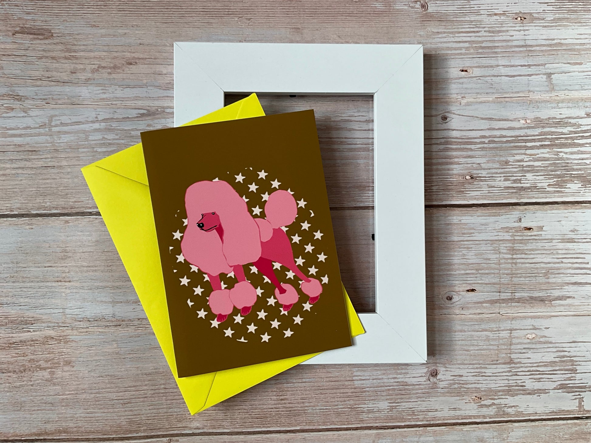 Greeting card with pink poodle design on a dark mustard background, yellow envelope, and white frame on wooden surface