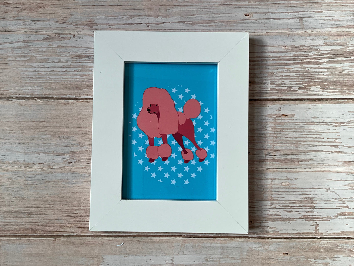 Framed mini print artwork of a pink poodle on a blue background with white stars, placed on a wooden surface.