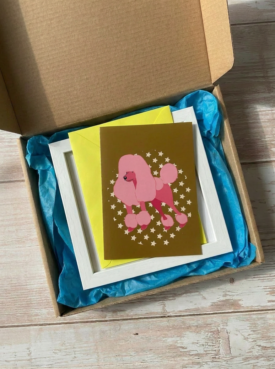 Pink poodle illustration on a greetings card with a fun yellow envelope inside a card mailing box with a white wooden frame to frame the card.