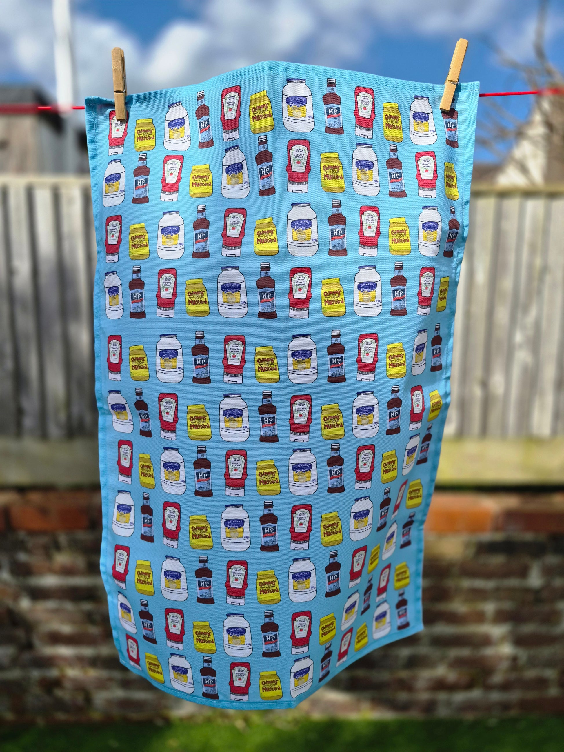 Unique blue tea towel with illustrated condiments print hanging on a washing line. 