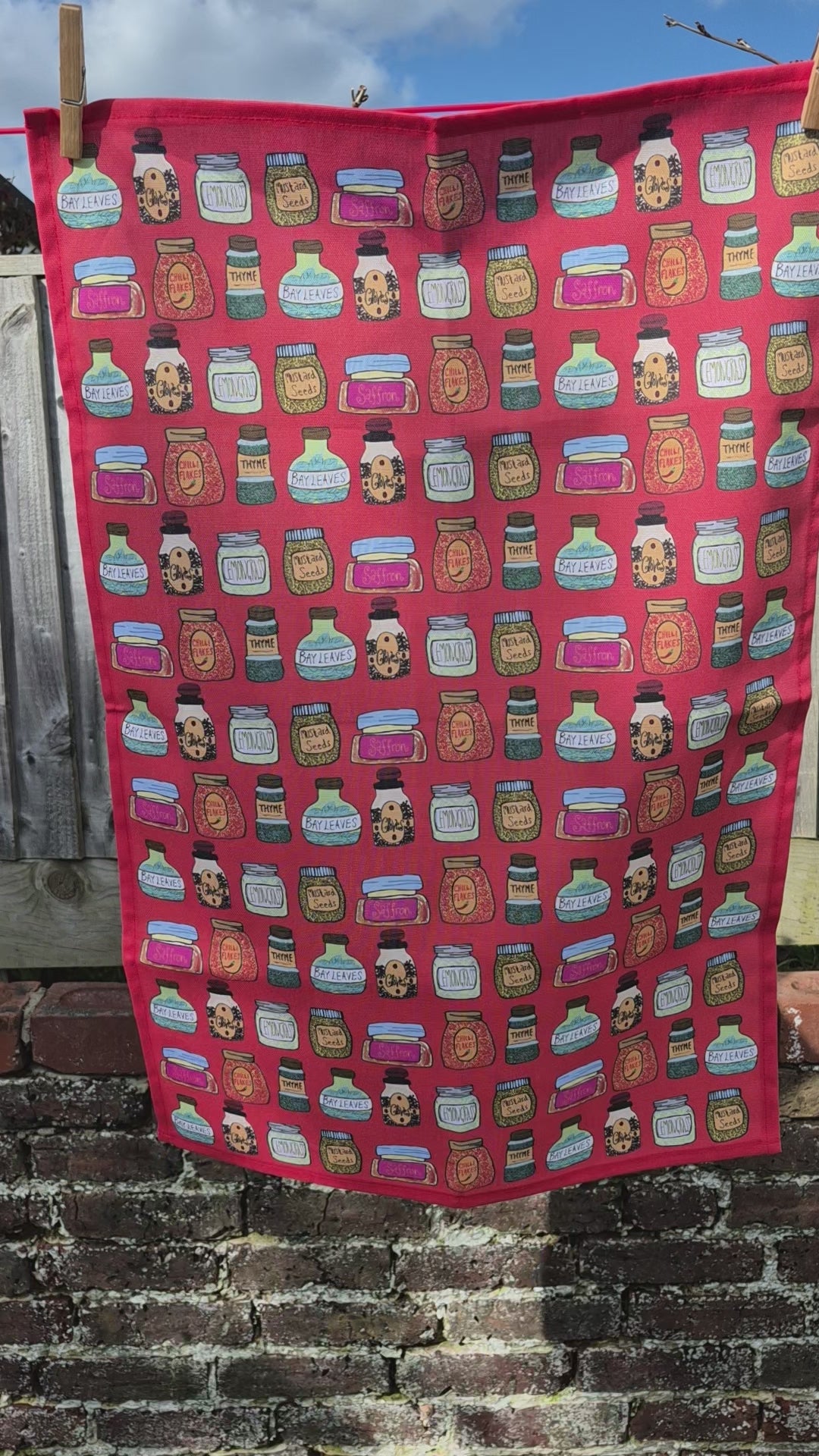 A video of a red tea towel with a illustrated printed design hanging on a washing line flowing in the breeze