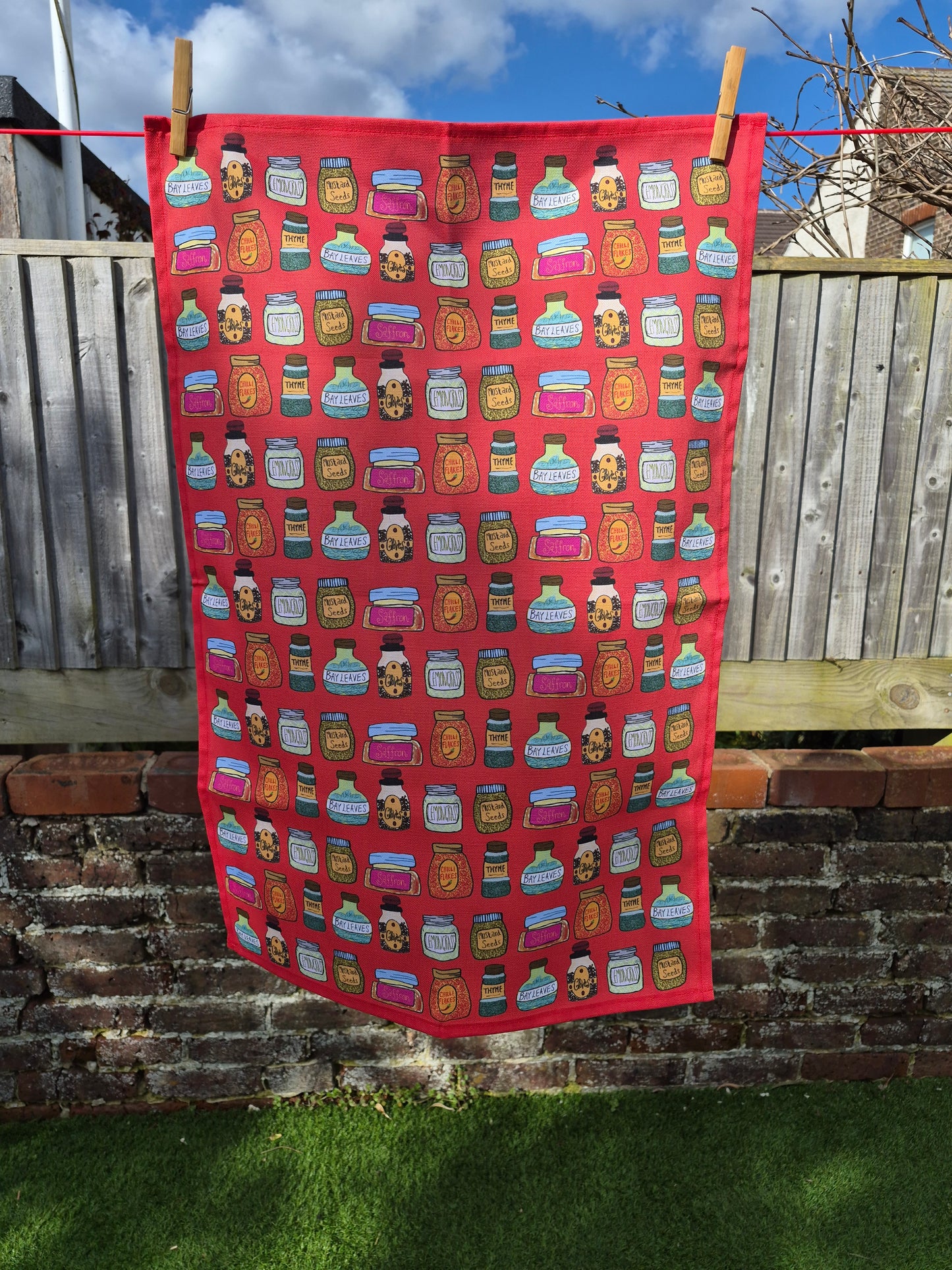 A red cotton tea towel with hand-drawn print of spice bottles hanging on a washing line