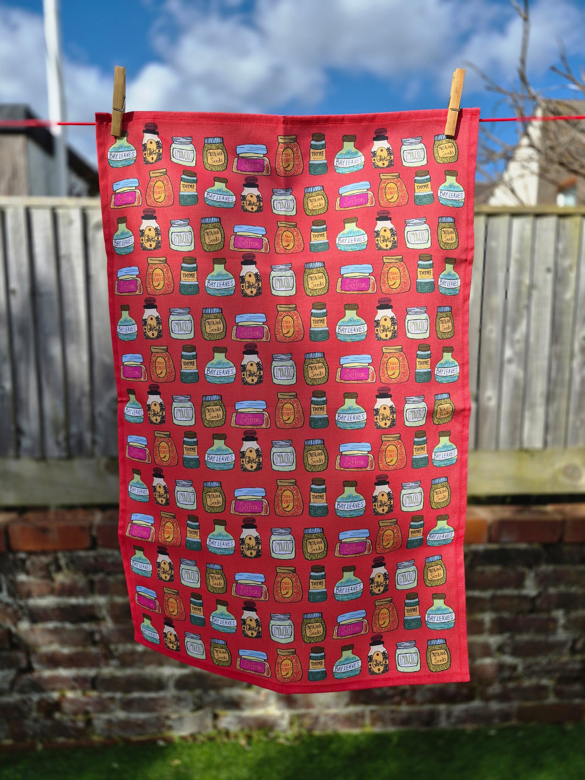 A product photo of a red cotton printed tea towel hanging on a washing line